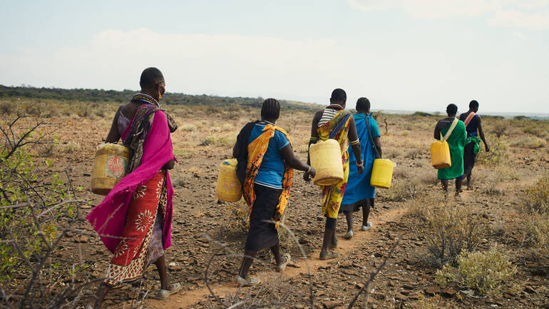 Kenianische Frauen auf dem Weg zu einem Wasserloch Sechs Frauen aus Kenia sind mit Kanistern auf dem Weg durch eine karge Landschaft, um Wasser an einem Wasserloch zu holen.