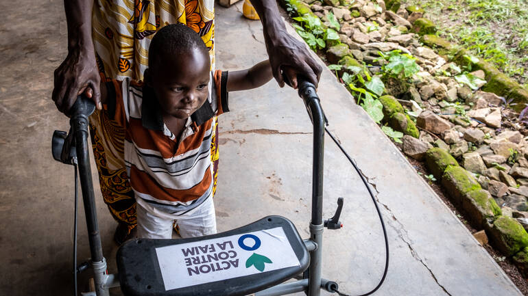 Dieubéni schafft mithilfe eines Rollators von Aktion gegen den Hunger und der Unterstützung seiner Großmutter schon erste Schritte über den Gehweg im Innenhof des Rehazentrums.
