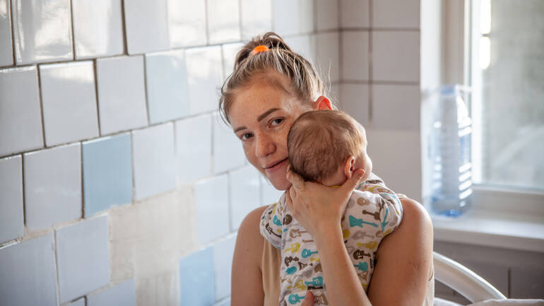 Eine junge Mutter in einem Kinderkrankenhaus in Moldau hält ihr Neugeborenes schützend im Arm und lächelt in die Kamera. Eine junge Mutter in einem Kinderkrankenhaus in Moldau hält ihr Neugeborenes schützend im Arm und lächelt in die Kamera.