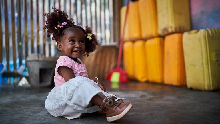 Mariame sitzt auf dem Boden vor Kanistern und lacht fröhlich. Mariame sitzt auf dem Boden vor Kanistern und lacht fröhlich.