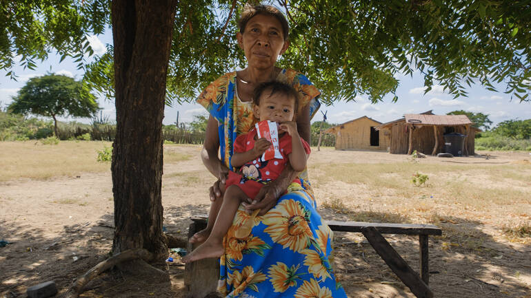 Eine ältere Frau sitzt mit einem kleinen Kind auf dem Schoß auf einer Bank. Das Kind hat eine Tüte Erdnusspaste in der Hand. Eine ältere Frau sitzt mit einem kleinen Kind auf dem Schoß auf einer Bank. Das Kind hat eine Tüte Erdnusspaste in der Hand.