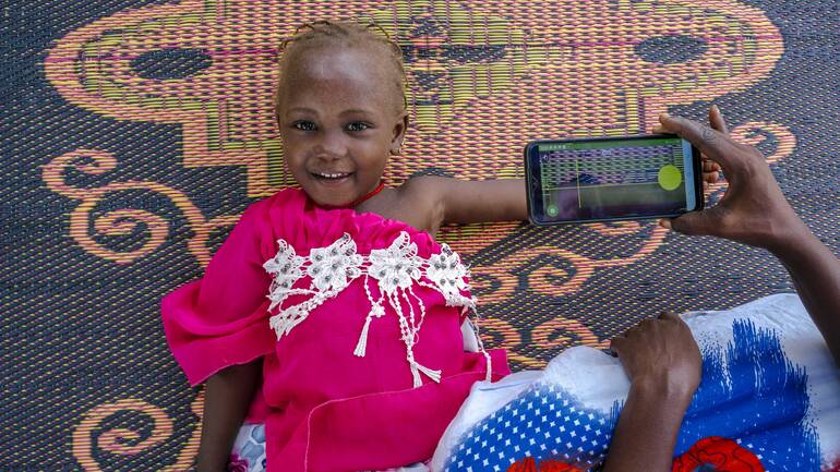 Ein Mädchen liegt lächelnd auf einem Teppich, während eine Frau ihren Arm mithilfe der SAM Photo App fotografiert. Ein Mädchen liegt lächelnd auf einem Teppich, während eine Frau ihren Arm mithilfe der SAM Photo App fotografiert.