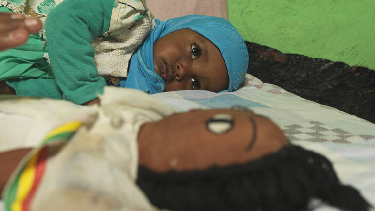 Munira liegt im Bett, im Vordergrund ihre Puppe.