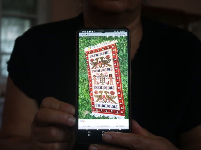 Eine Person hält den Bildschirm eines Smartphones in die Kamera, auf dem ein handgewebter Teppich zu sehen ist.