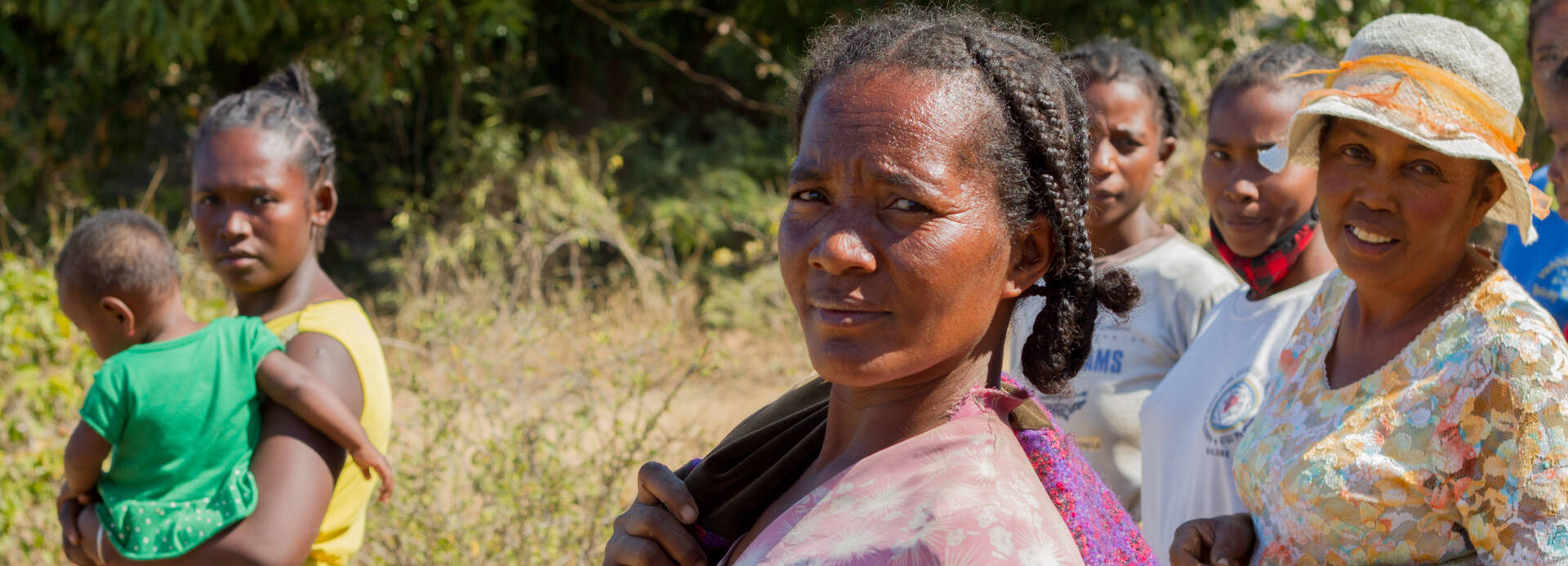 Frauen aus Madagaskar auf dem Weg zum Garten Mehrere Frauen sind auf dem Weg zum Schulgarten, eine trägt ein Kind auf dem Arm, sie gucken in die Kamera, im Hintergrund trockene Umgebung.