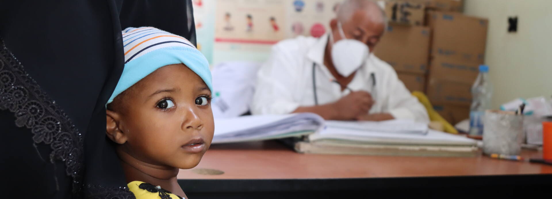 Ein Kind in den Armen der Mutter schaut in die Kamera, im Hintergrund ein Arzt aus dem Gesundheitszentrum in Almakhzan im Jemen.