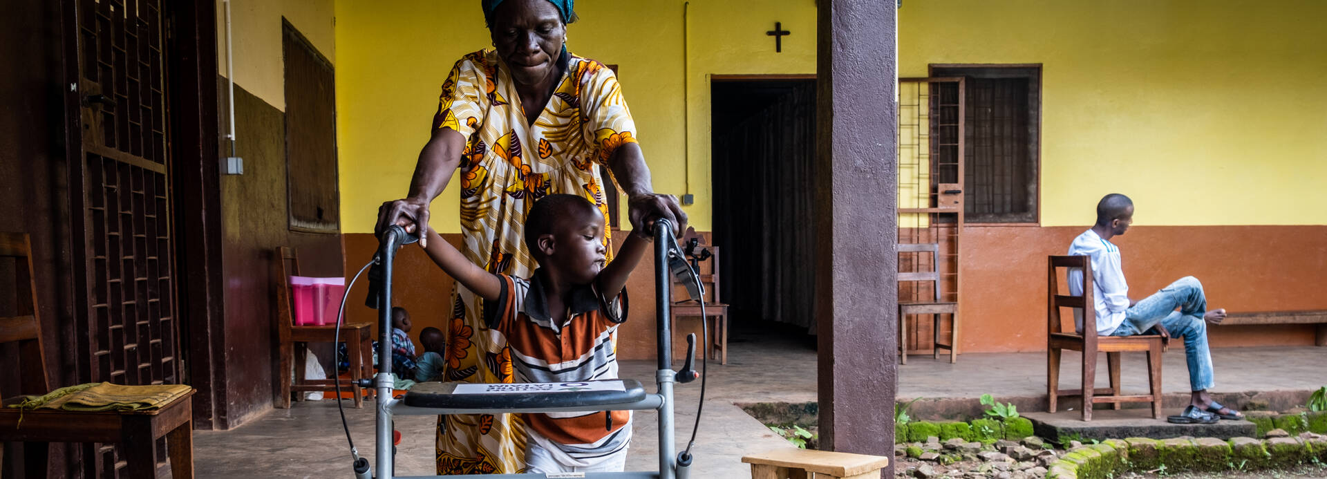 Dieubéni kann mithilfe eines Rollators endlich das Laufen lernen. Seine Großmutter stützt ihn dabei und hält seine Hände am Gehwagen fest, während sie durch den Innenhof des Therapiezentrums laufen. 