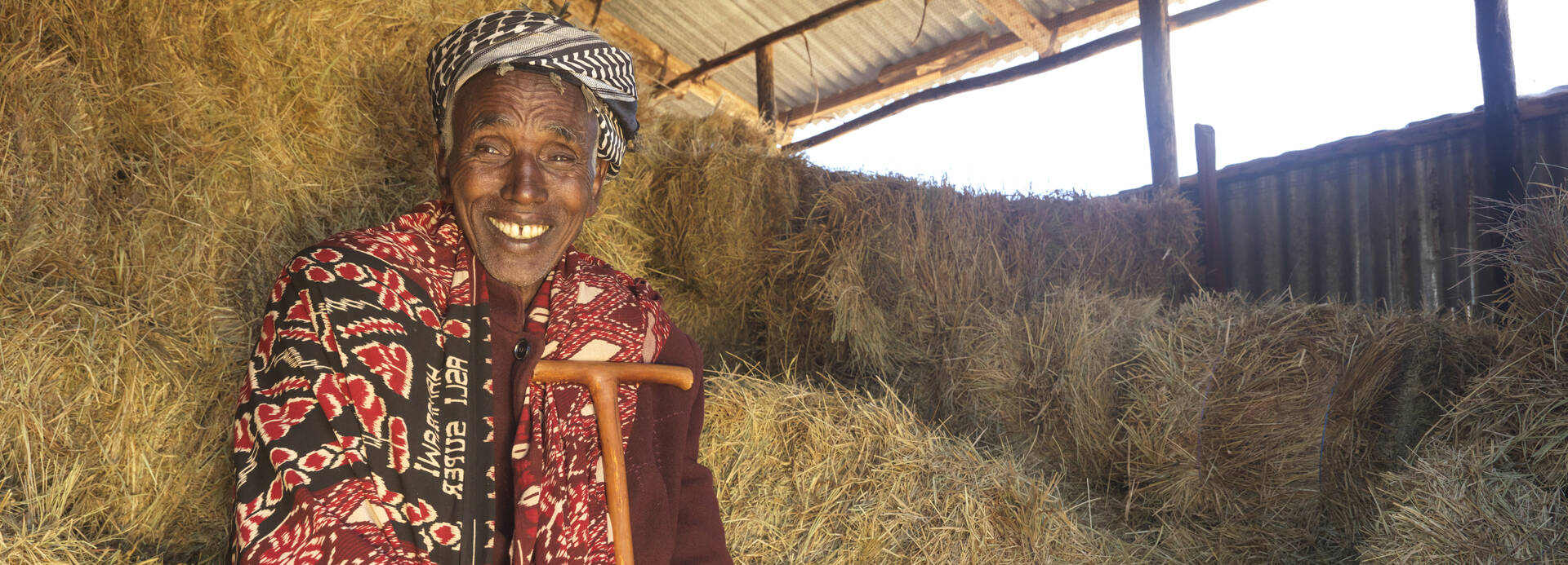 Elimule sitzt lachend im Lagerhaus zwischen Heuballen, die sein Vieh ernähren und seiner Familie ein neues Leben ermöglichen. 