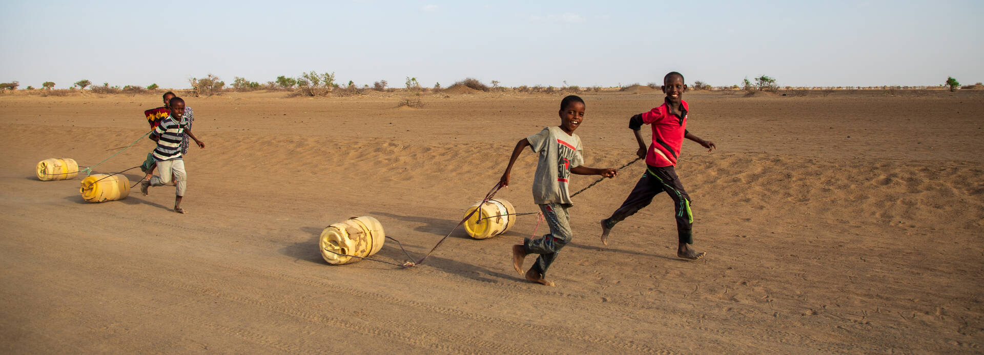 Vier Jungs aus Kenia laufen mit Wassertanks an Seilen über einen Wüstenweg. Vier Jungs aus Kenia laufen mit Wassertanks an Seilen über einen Wüstenweg.