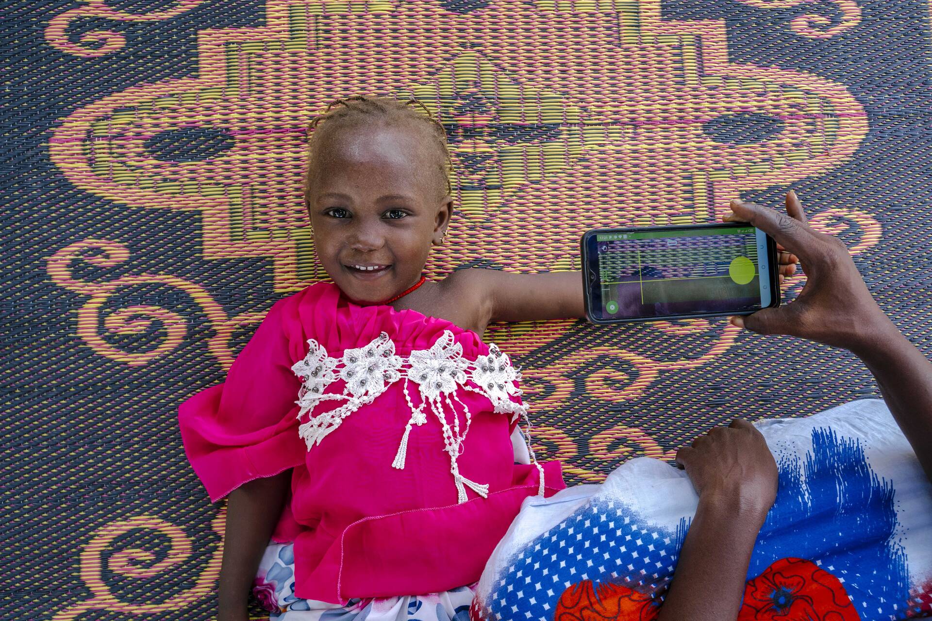 Ein Mädchen liegt lächelnd auf einem Teppich, während eine Frau ihren Arm mithilfe der SAM Photo App fotografiert.