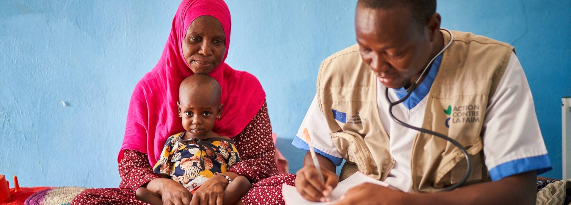 Ein Arzt von Aktion gegen den Hunger im Tschad bei der Anamnese Eine Frau sitzt mit ihrem Kleinkind auf einer Liege. Rechts neben ihnen sitzt ein Arzt von Aktion gegen den Hunger und trägt etwas in ein Formular ein.