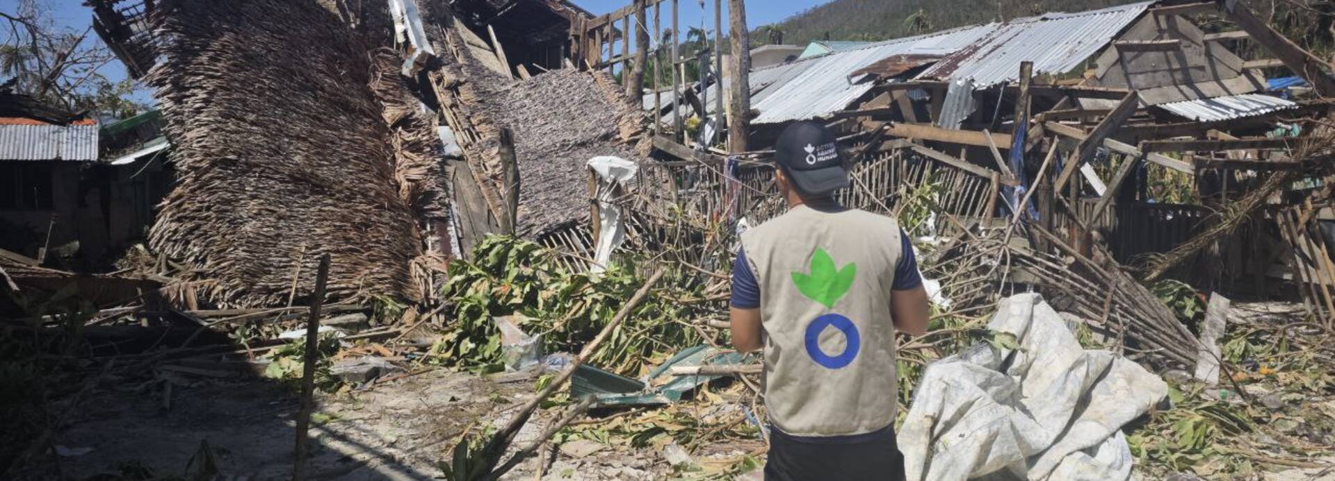 Mitarbeiter von Aktion gegen den Hunger vor zerstörten Häusern durch Taifun in Barangay Liberty, Hibisong Island, Loreto Dinagat Islands (Philippinen)