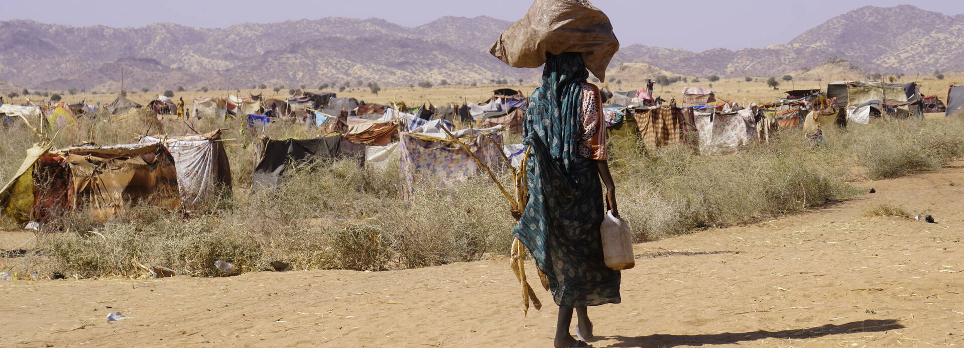 Eine Person im Sudan mit Gepäck auf dem Kopf geht durch eine trockene Landschaft, im Hintergrund ist ein provisorisches Lager zu sehen
