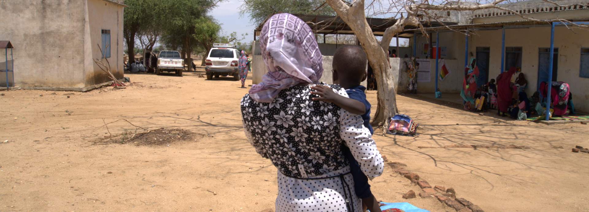 Mutter mit Kind in der Rückansicht. Die Fraz erhält in einer mobilen Klinik im Sudan therapeutische Spezialnahrung (RUTF) und Medikamente für ihr Kind.