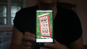 Eine Person hält den Bildschirm eines Smartphones in die Kamera, auf dem ein handgewebter Teppich zu sehen ist.
