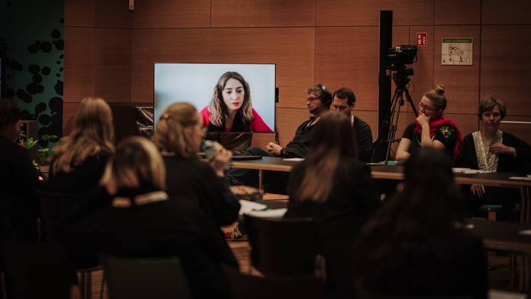 Eine dazugeschaltete Teilnehmerin auf dem Bildschirm gibt Input, die Menschen im Raum hören zu. Eine dazugeschaltete Teilnehmerin auf dem Bildschirm gibt Input, die Menschen im Raum hören zu.