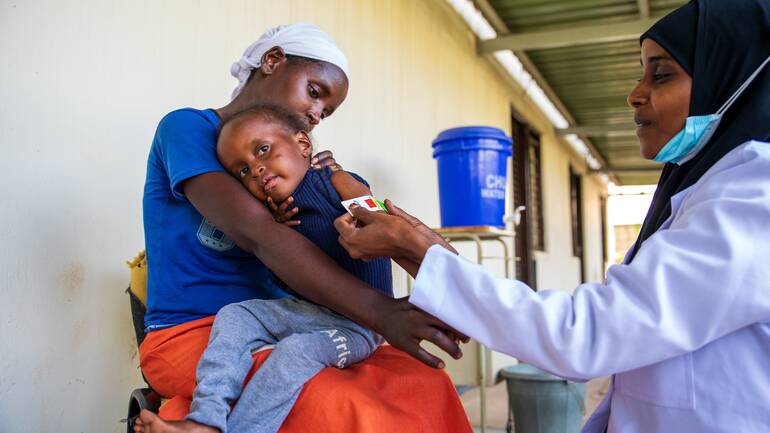 Khadija, ein Mädchen aus Kenia, wird auf dem Schoß ihrer Mutter sitzend von einer Krankenschwester mit einem MUAC-Band untersucht.