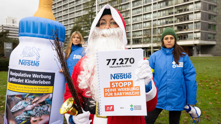 77.270 Unterschriften hat der Nikolaus zusammen mit Aktion gegen den Hunger für Nestlé mitgebracht - der Konzern bekommt dieses Jahr die Rute, denn den Milchkodex der WHO halten sie noch immer nicht ein. Der Nikolaus hält ein Buch mit den 77.270 Unterschriften gegen Nestlés Werbestrategie hoch, im Hintergrund eine große Babyflasche und zwei Mitarbeiterinnen von Aktion gegen den Hunger - alle vor dem Nestlé-Hauptsitz in Frankfurt am Main.