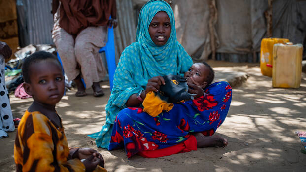 Eine junge somalische Mutter sitzt mit ihrem Baby auf dem Boden, im Vordergrund schaut ein Junge in die Kamera.