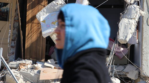 Das Ausmaß der Zerstörung nach den Erdbeben in der Türkei und in Syrien ist für die Menschen kaum fassbar. Vorne eine Person mit blauer Kapuze, unscharf, dahinter ein Teil eines zusammengestürzten Hauses, scharf