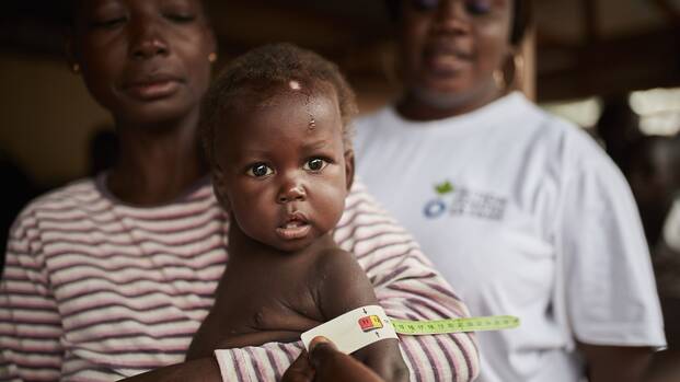 Ein Kind mit wird mit MUAC-Band auf Mangelernährung untersucht, während die Mutter es im Arm hält, im Hintergrund eine Mitarbeiterin von Aktion gegen den Hunger.