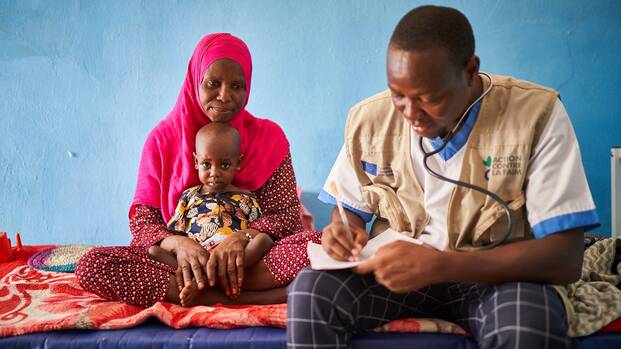 Ein Arzt von Aktion gegen den Hunger im Tschad bei der Anamnese Eine Frau sitzt mit ihrem Kleinkind auf einer Liege. Rechts neben ihnen sitzt ein Arzt von Aktion gegen den Hunger und trägt etwas in ein Formular ein.