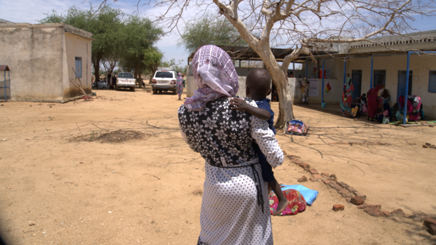 Mutter mit Kind in der Rückansicht. Die Fraz erhält in einer mobilen Klinik im Sudan therapeutische Spezialnahrung (RUTF) und Medikamente für ihr Kind.